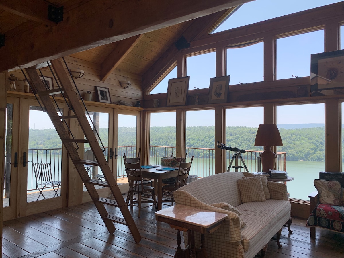 A bright and inviting living area features large windows that provide views of the surrounding landscape. A ladder leads to a loft space, while a comfortable couch and a wooden dining table create a cozy atmosphere. Natural light enhances the wooden accents throughout the room.