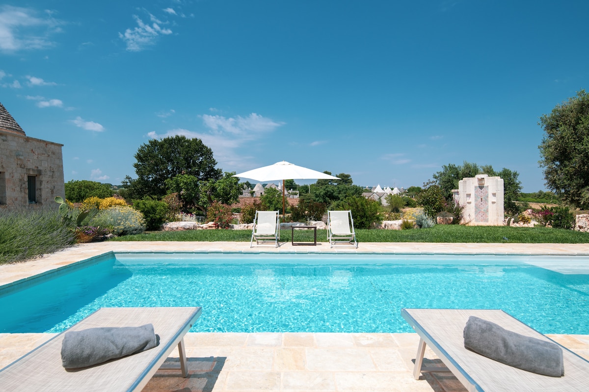 A tranquil salt-water pool is surrounded by lush greenery, with two sun loungers positioned at the pool's edge. A large umbrella provides shade, while vibrant flowers and trees frame the serene landscape under a clear blue sky.