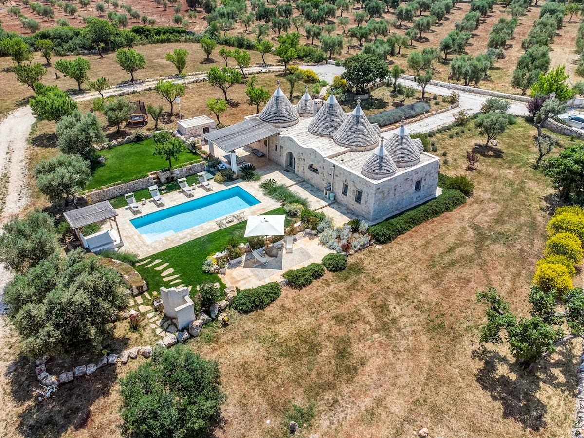 Aerial view of the villa showcases its distinctive trulli architecture surrounded by lush greenery. The spacious outdoor area includes a generously sized salt-water pool and shaded seating areas, with a beautifully landscaped garden featuring rows of young olive trees in the background.