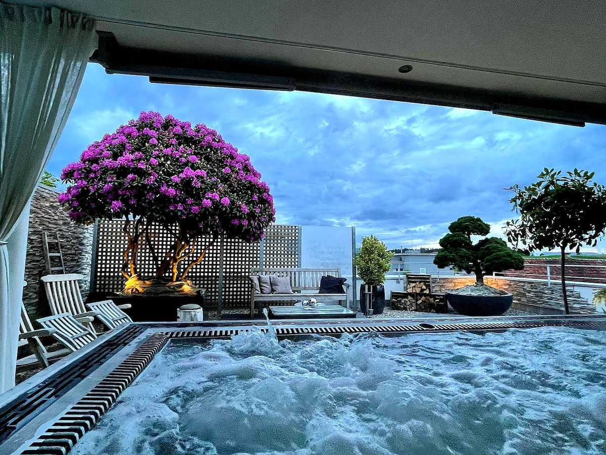 An outdoor area features a hot jacuzzi surrounded by relaxing seating and vibrant flowers in pots. A large bougainvillea tree adds color against a backdrop of cloudy skies. Soft lighting enhances the tranquil atmosphere, inviting guests to unwind in a serene setting.
