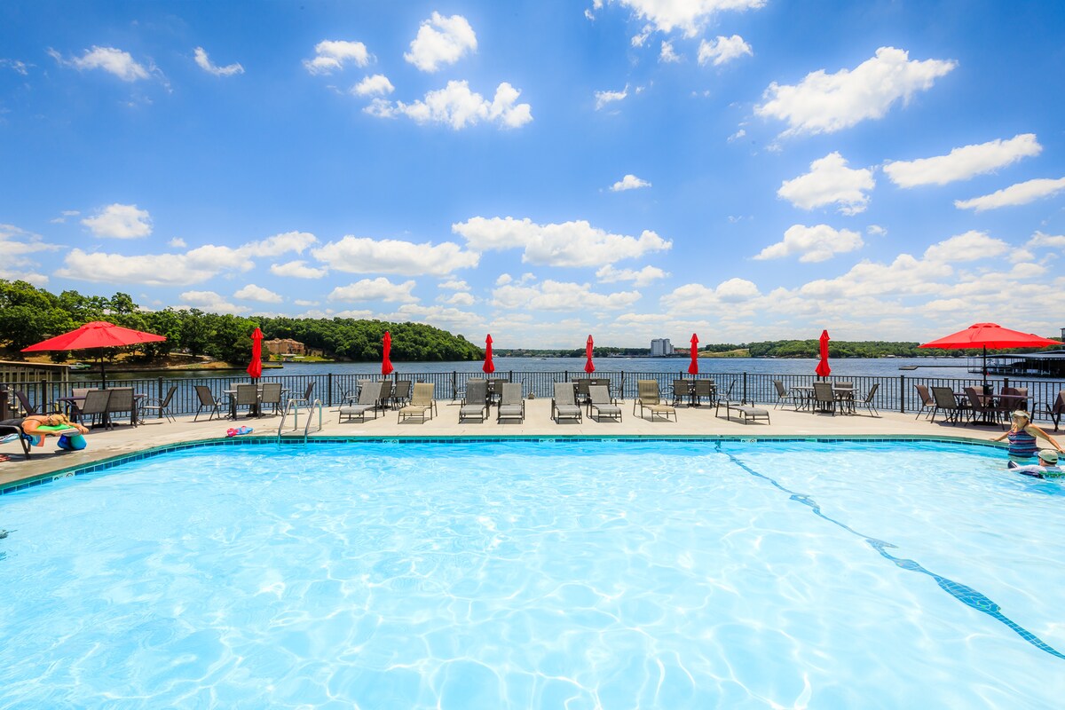 A swimming pool is centered in the foreground, featuring clear blue water and surrounded by lounge chairs. Red umbrellas provide shade along the poolside, overlooking a scenic lake view under a bright sky filled with fluffy clouds.