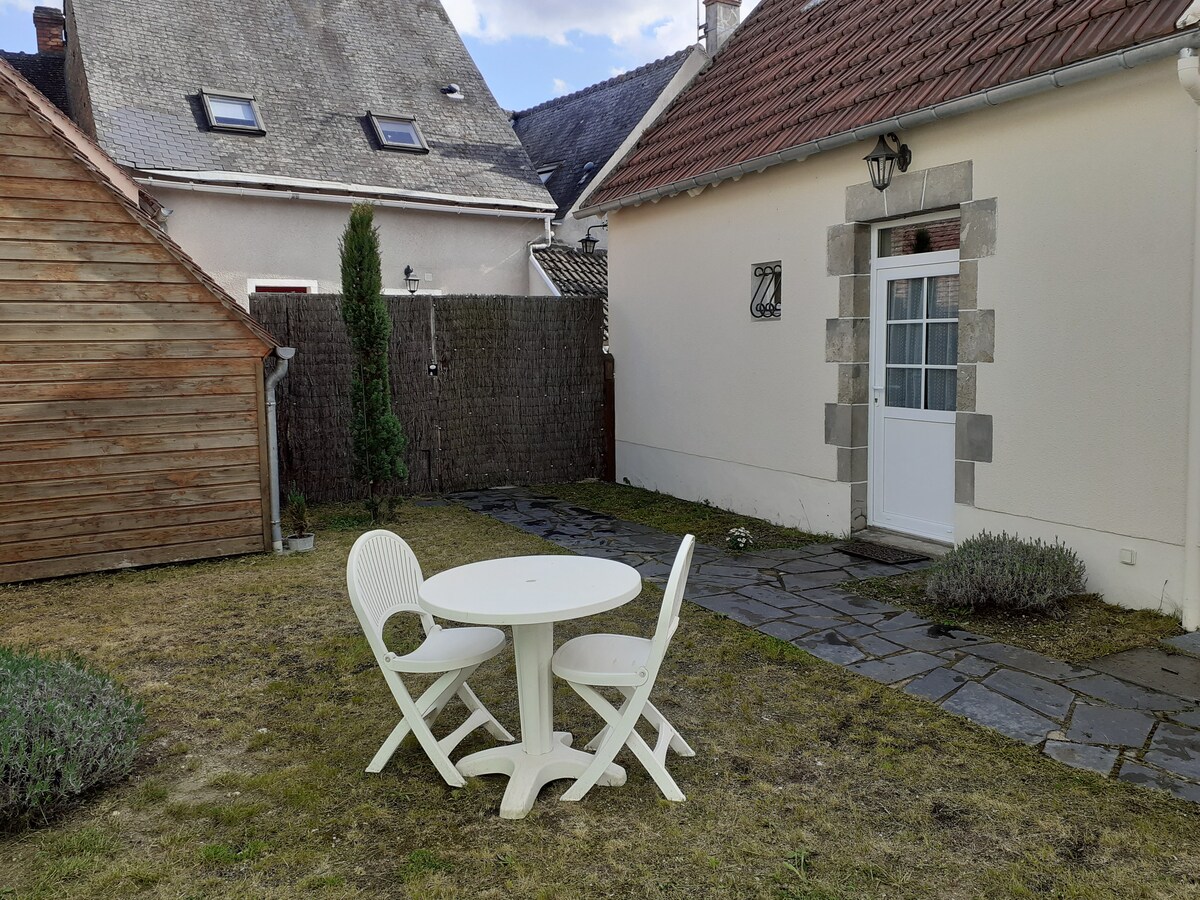 A private courtyard is featured with a round white table surrounded by two white chairs. Natural grass covers the ground, and a low wooden fence borders the yard, providing a sense of privacy. Nearby, a small building is partially visible.