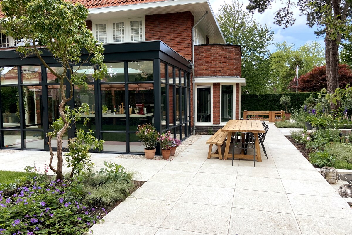 A spacious outdoor area features a long wooden table surrounded by black chairs, set on a stone patio. Lush greenery and potted flowers enhance the space, while a modern glass extension of the house offers views of the interior. Trees provide gentle shade.