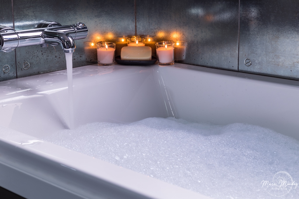 A modern bathtub is filled with foam, with water cascading from a sleek chrome faucet. Candles are positioned around the tub, providing gentle illumination in a serene setting. A soap bar rests on the ledge, enhancing the relaxing ambiance.