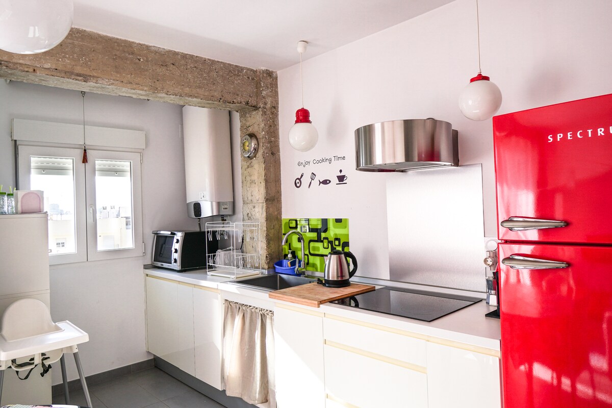 A modern kitchen is equipped with a red refrigerator and a sleek cooking range. Green backsplash tiles add a vibrant touch. Natural light enters through two windows, illuminating the space. Countertops are clear, offering ample preparation area. A kettle and a cutting board are positioned on the stove.