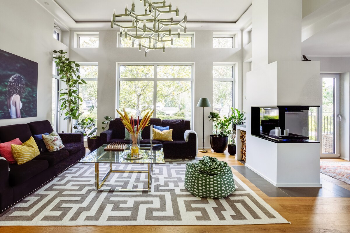 A spacious living area is highlighted by large windows allowing natural light to fill the space. Comfortable seating is arranged around a glass coffee table on a patterned area rug, with greenery adding freshness. A modern fireplace complements the contemporary decor.