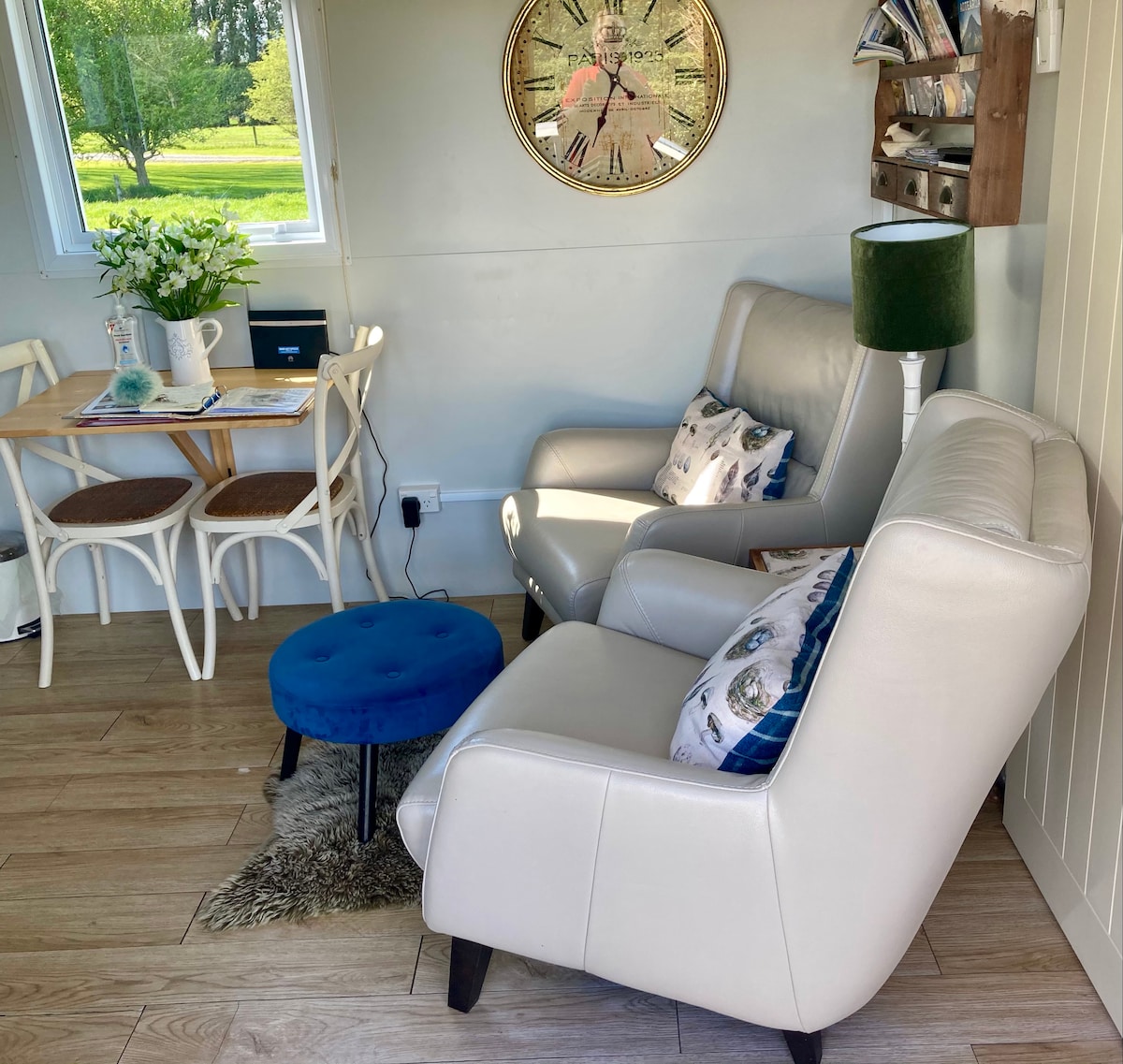 A cozy corner features a light-colored armchair and a matching footstool, complemented by a decorative pillow. A small wooden table with white chairs is set nearby, adorned with a vase of flowers and a magazine rack on the wall. Natural light floods in through a window, enhancing the inviting space.