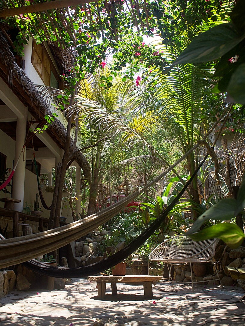 A serene garden space is surrounded by tall tropical plants and trees. A hammock is gently draped between two trunks, providing a cozy resting area. A small table made of wood is positioned on the ground, adding to the natural ambiance of the retreat.