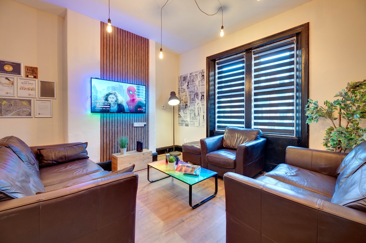 A modern living room features two brown leather sofas arranged around a glass coffee table. A wall-mounted TV, positioned above a decorative console, displays vibrant content. Large windows with horizontal blinds let in natural light, complemented by a floor lamp and potted plant in the corner.