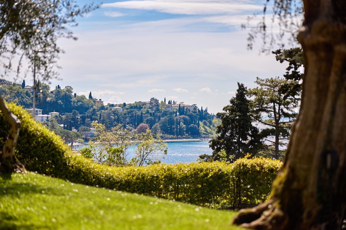 A scenic view of Lake Garda is captured, showcasing tranquil waters framed by lush greenery and trees. The landscape features distant hills dotted with buildings and vegetation, creating a peaceful backdrop against the clear sky.