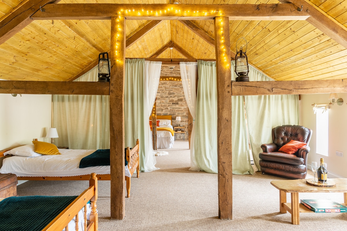 An open and spacious loft area features high wooden ceilings with exposed beams. Two beds, including a bespoke large bed, are visible, separated by sheer curtains. Soft lighting is provided by lanterns, and a cozy armchair is placed next to a small coffee table.