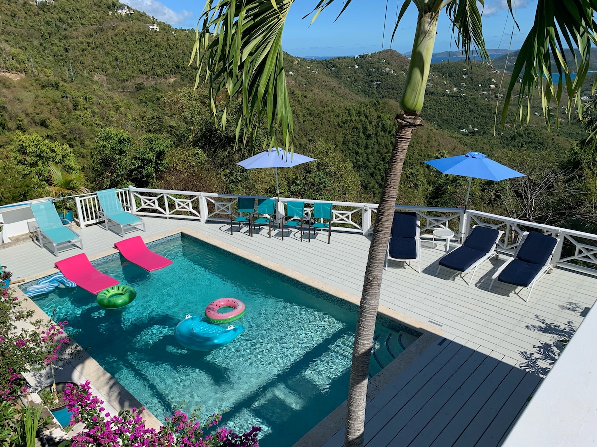 A well-maintained swimming pool is surrounded by a spacious deck featuring lounging chairs in various colors. Umbrellas provide shade, while a variety of pool floats rest on the water. Lush greenery borders the area, enhancing the serene environment.