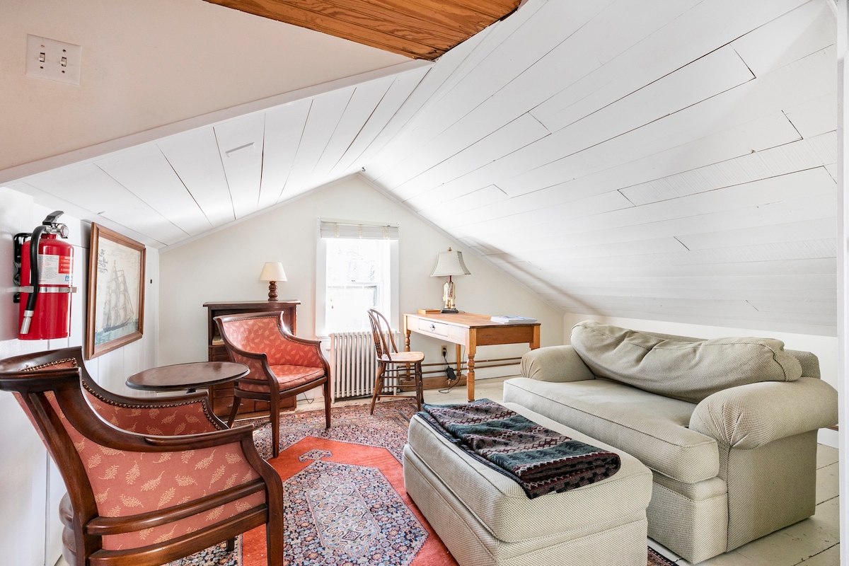 A cozy loft area is enhanced by a sloped ceiling, featuring a comfortable sofa, two armchairs, and a small round table. A writing desk is positioned by a window, offering natural light. A fire extinguisher is securely mounted on the wall for safety.