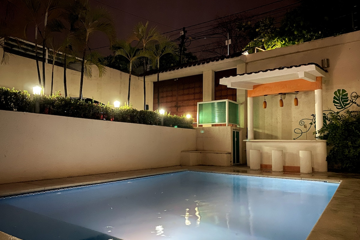 An inviting pool area is highlighted by tranquil water under soft evening lighting. Lush landscaping surrounds the space, complemented by a small bar section with light fixtures. The overall ambiance offers a peaceful environment for relaxation.