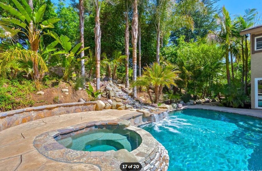 A tranquil outdoor space showcases a sparkling pool with a waterfall feature, surrounded by lush tropical landscaping. A stone hot tub is positioned nearby, inviting relaxation. The scene is enhanced by palm trees and greenery, creating a serene retreat.