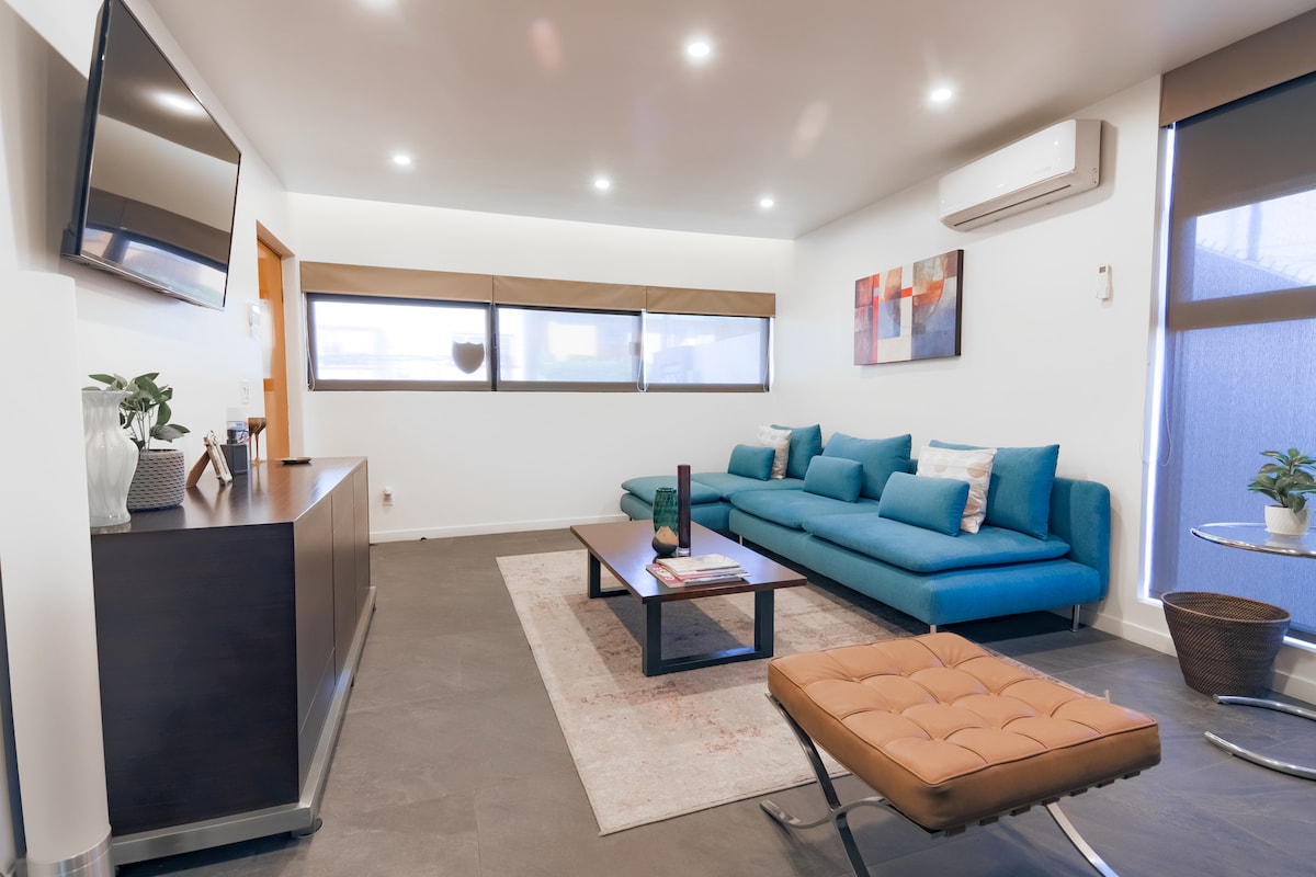 A modern living room features a large blue sectional sofa adorned with several cushions. A low coffee table occupies the center, complemented by a side table with a sleek design. Natural light enters through the large windows, drawing attention to the minimalist decor and contemporary furnishings.