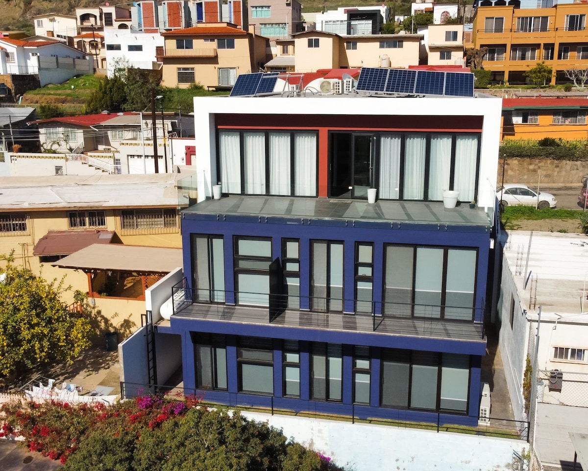 The modern building features multiple levels with a sleek facade that combines dark blue and white elements. Large glass windows and sliding doors allow natural light to fill the interiors. A rooftop terrace with solar panels adds to the building's contemporary design.