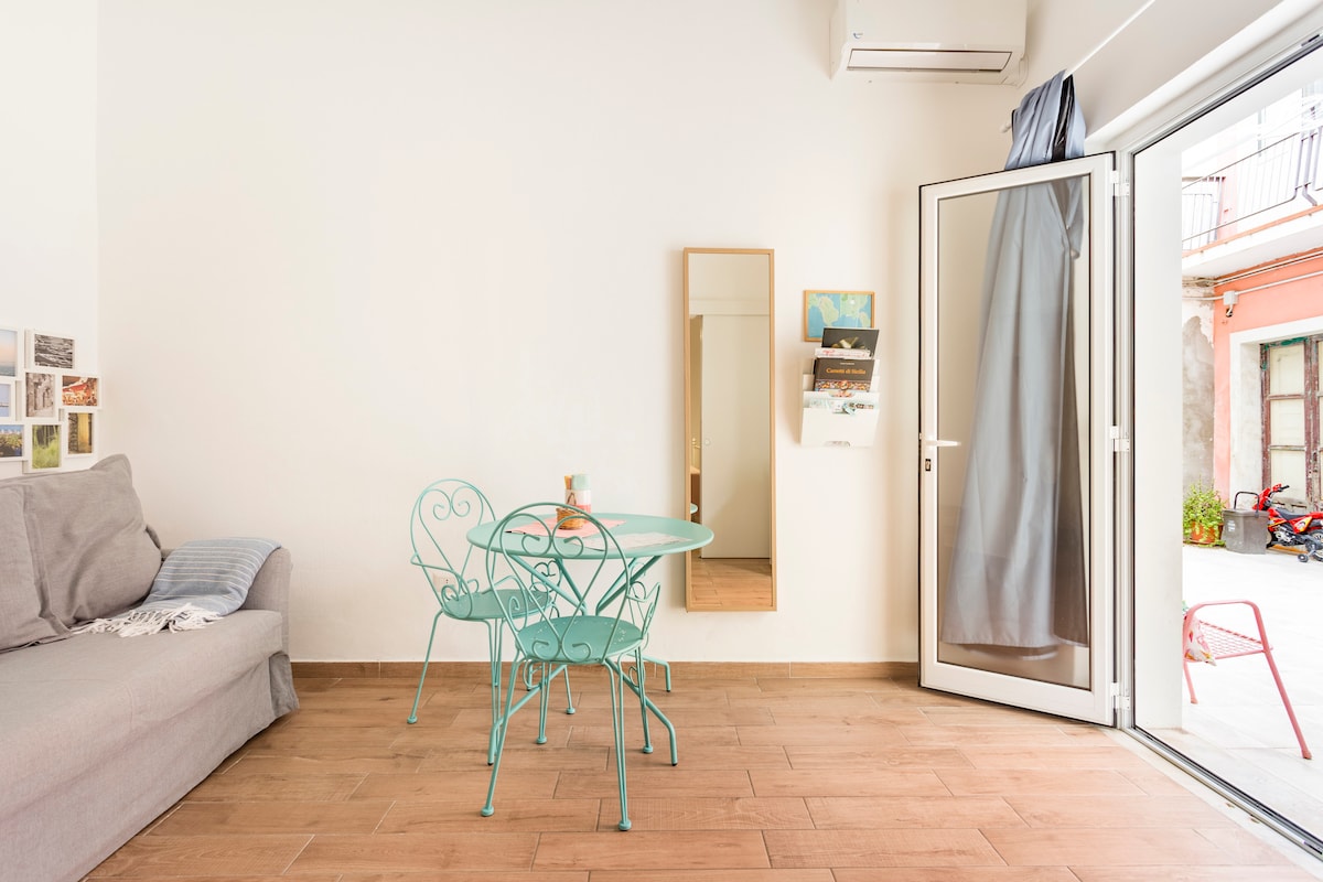 A light-filled living area is complemented by wooden flooring and a cozy gray sofa. A round turquoise table with two matching chairs invites casual dining. A large mirror reflects the space, and a door opens directly to the quiet courtyard, enhancing the room's brightness.
