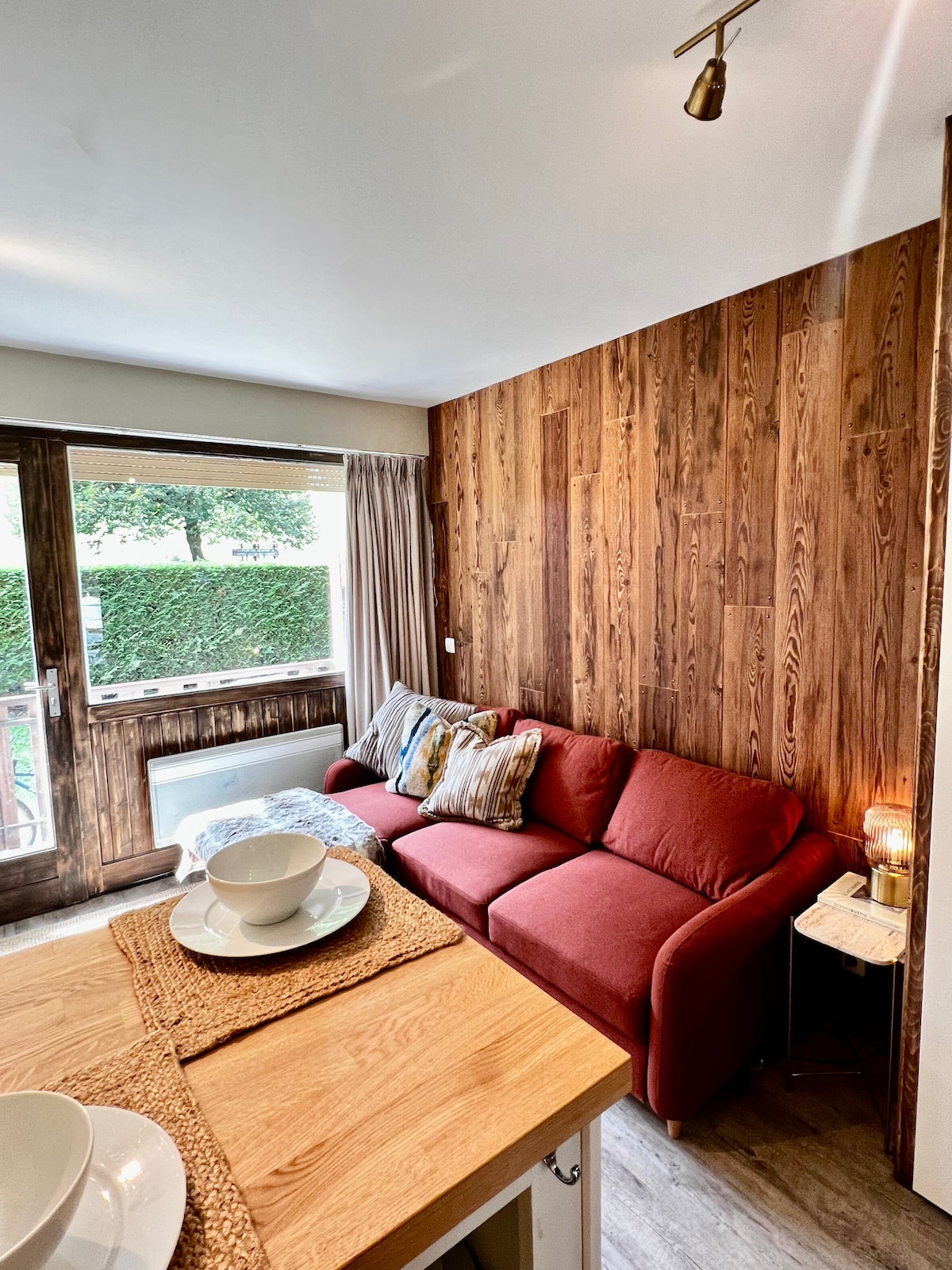 A cozy living space showcases a comfortable red sofa, complemented by decorative cushions. Warm wooden walls add a rustic touch, while natural light filters through the window, illuminating the area. A wooden dining table is set nearby, ready for meals.