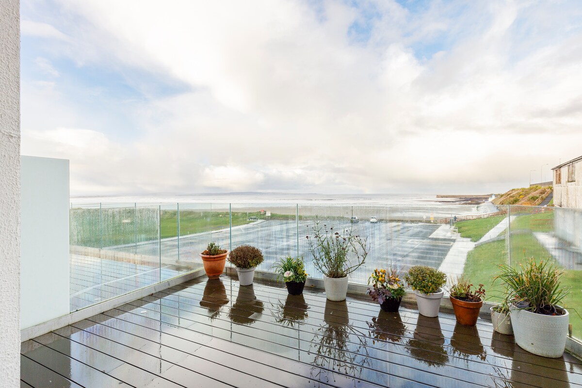 A spacious deck is featured, lined with potted plants, overlooking the beach and the Atlantic Ocean. The weathered wooden surface reflects the surrounding views, and the glass railing ensures unobstructed sightlines of the horizon. The scene is punctuated by soft, natural lighting.