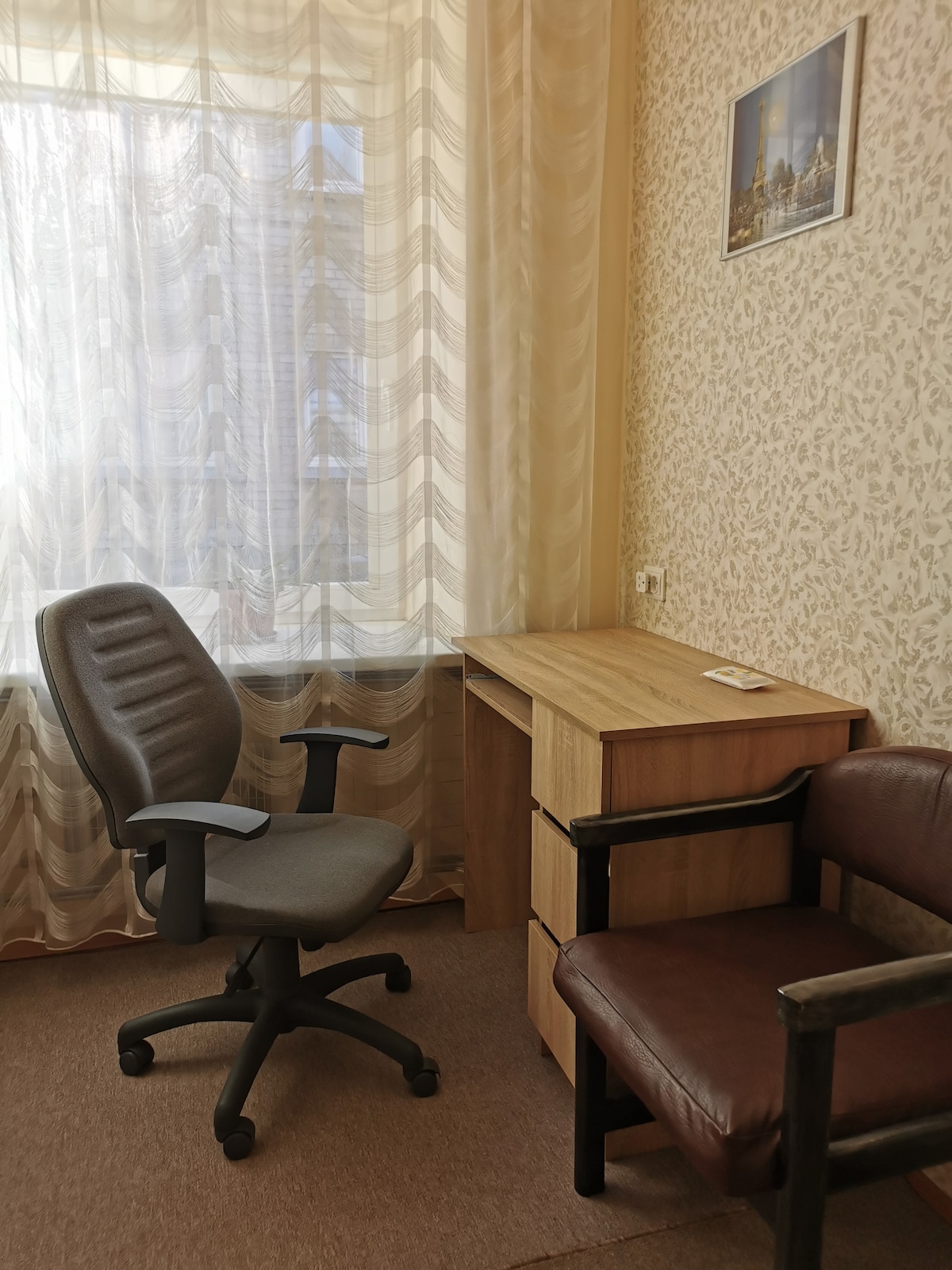 A workspace is featured with a wooden desk and a two-drawer unit. A rolling office chair sits beside the desk, and a brown armchair is positioned nearby. Soft curtains diffuse natural light from the window, highlighting the patterned wallpaper.