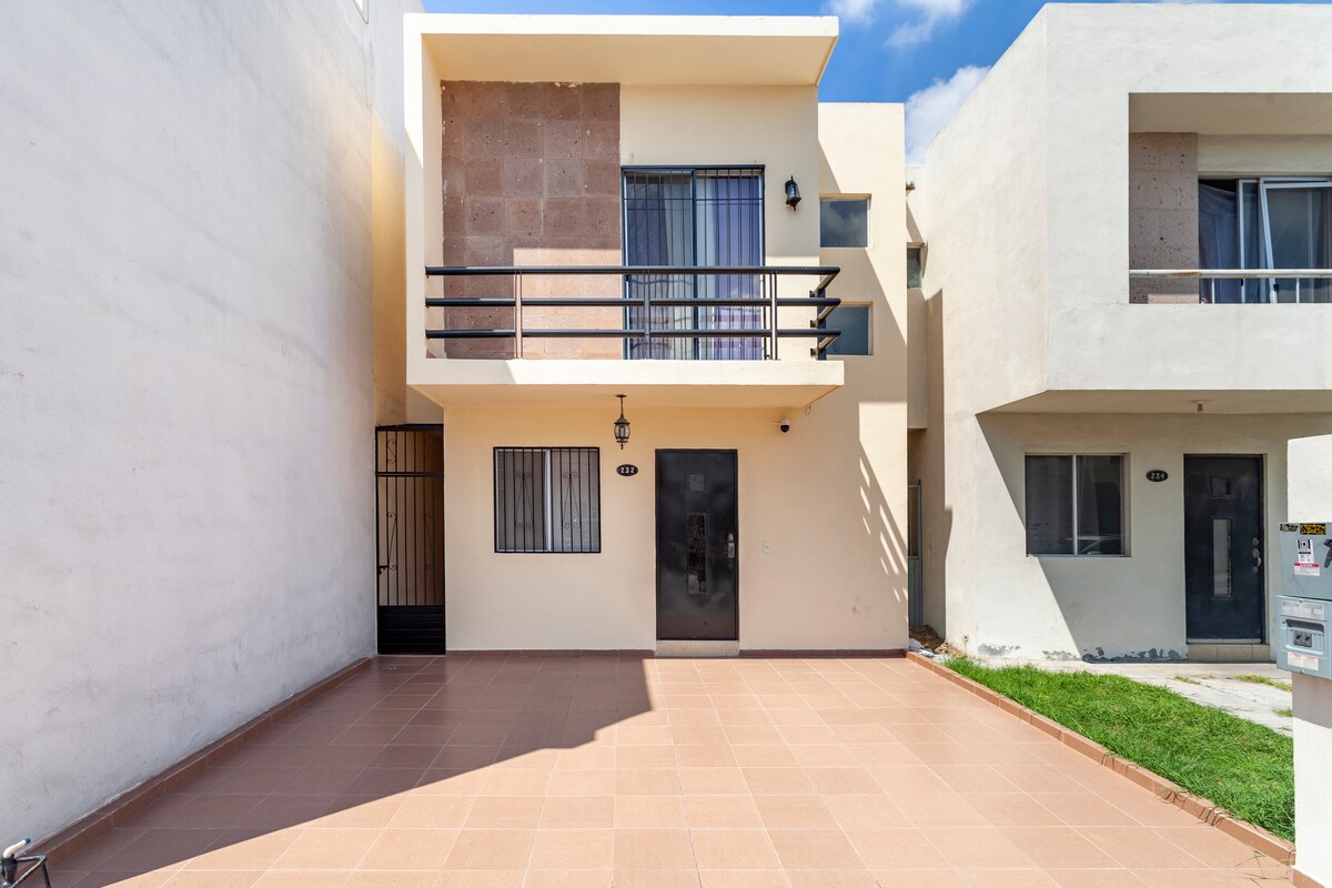 A modern two-story house is depicted, featuring a light-colored facade with stone accents. The entrance is framed by a black railing and a large window. An open patio area extends in front, showcasing a tiled surface and a patch of grass on the right.