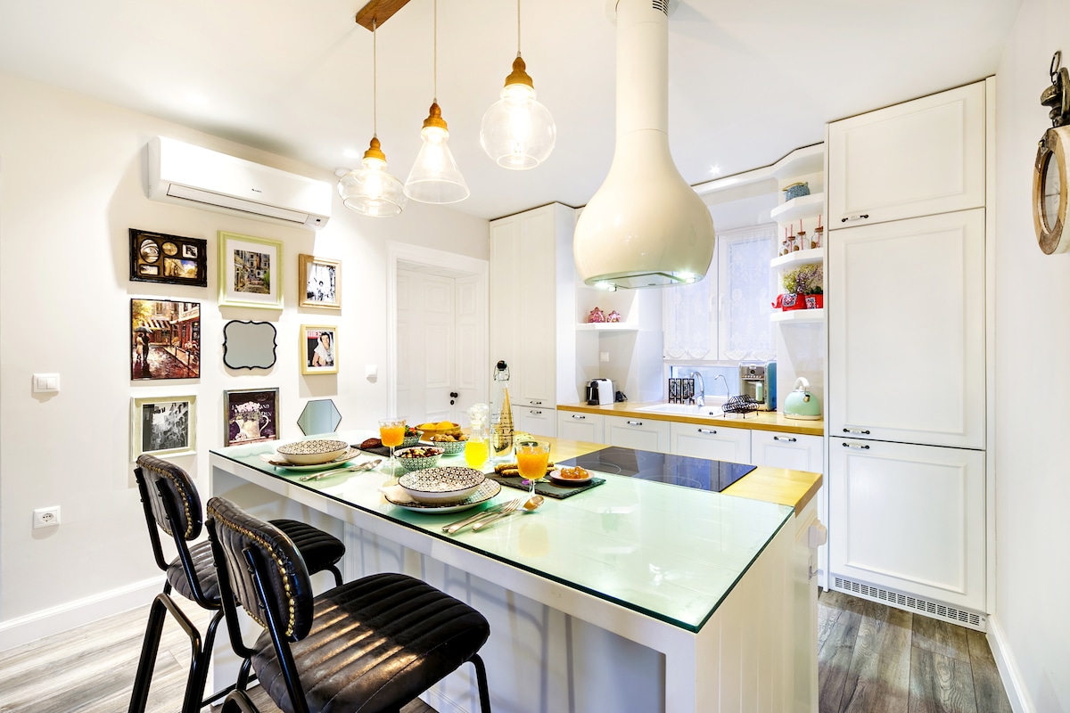A modern kitchen features a central island with a glass top and seating for four. Bright pendant lights hang overhead, illuminating the space. The walls display framed photographs, and essential appliances, including a kettle and coffee maker, are visible, offering functionality and style.