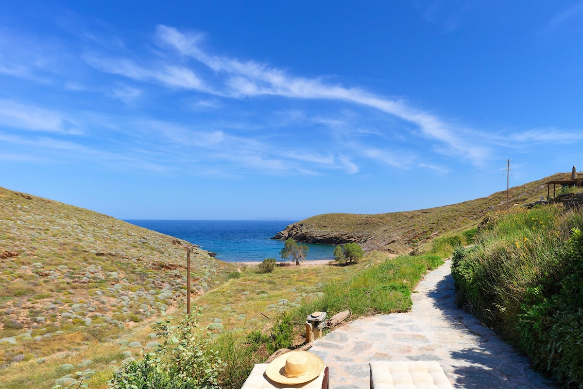 A scenic path leads through lush greenery towards a tranquil bay, with gentle hills framing the serene blue water. The expansive sky is dotted with wispy clouds, creating a peaceful backdrop for the idyllic landscape.