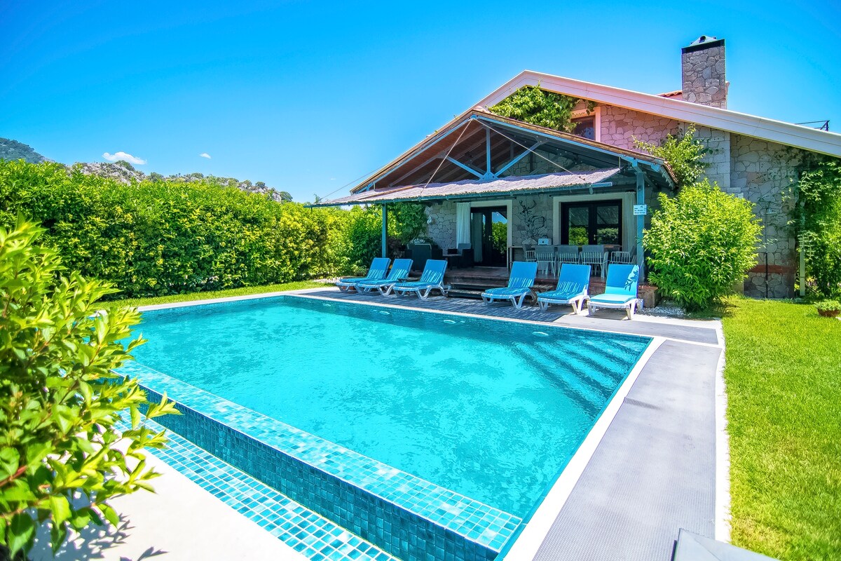 A private pool is surrounded by a landscaped garden, featuring a variety of green shrubs. Several lounge chairs are placed neatly along one side of the pool, under a shaded terrace of the villa. The building's stone and wood architecture complements the serene outdoor setting.