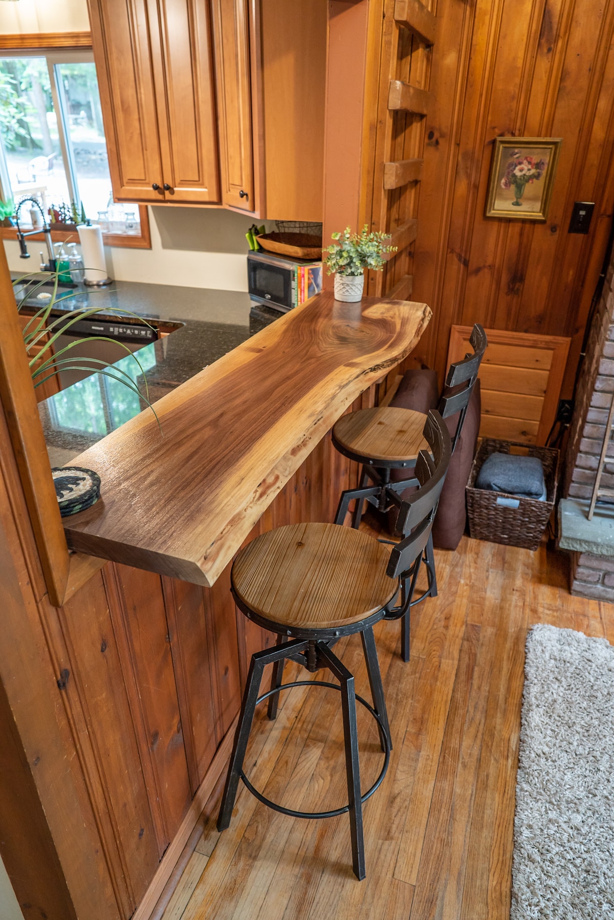A wooden countertop crafted from natural wood is situated next to a kitchen area, accompanied by three high stools. The warm, knotty pine walls complement the inviting atmosphere, while light enters through a nearby window illuminating the space.