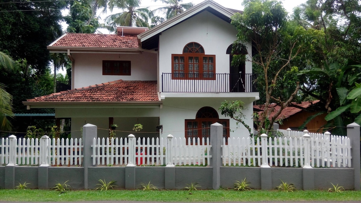 A two-story house is framed by a white picket fence, surrounded by lush greenery. The structure features a terracotta-tiled roof, large arched windows, and a balcony on the upper level. Tropical trees provide shade and contribute to the peaceful outdoor setting.