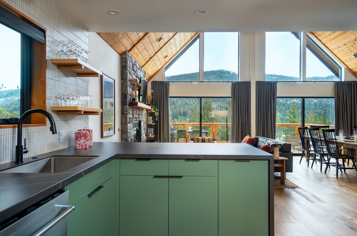 The kitchen features a green cabinetry design with a dark countertop, while large windows provide a view of the surrounding mountains. An open floor plan creates a spacious atmosphere, and the area is complemented by wood accents and carefully arranged seating in the adjacent living space.