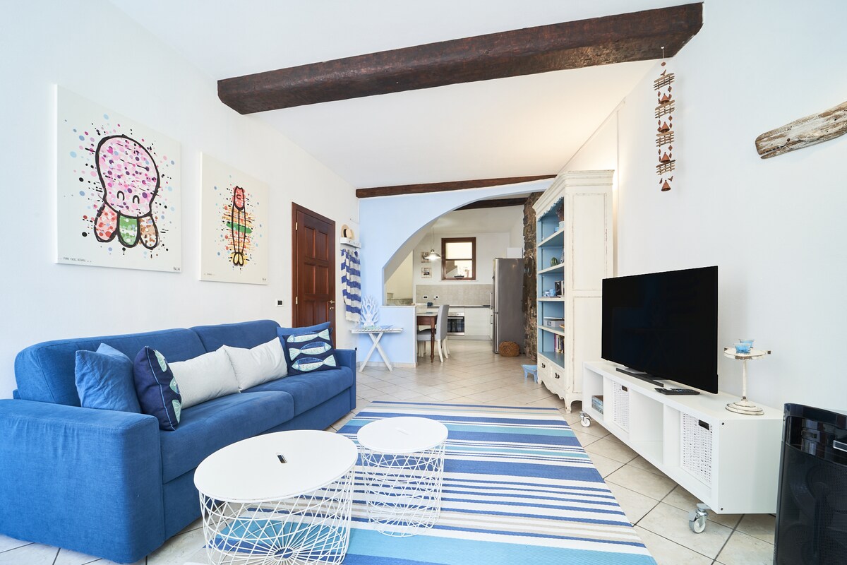 A spacious living area features a blue couch adorned with decorative pillows, accompanied by two white coffee tables. Colorful wall art enhances the atmosphere. A flat-screen television is positioned on a white media console, with an archway leading to the kitchen visible in the background.