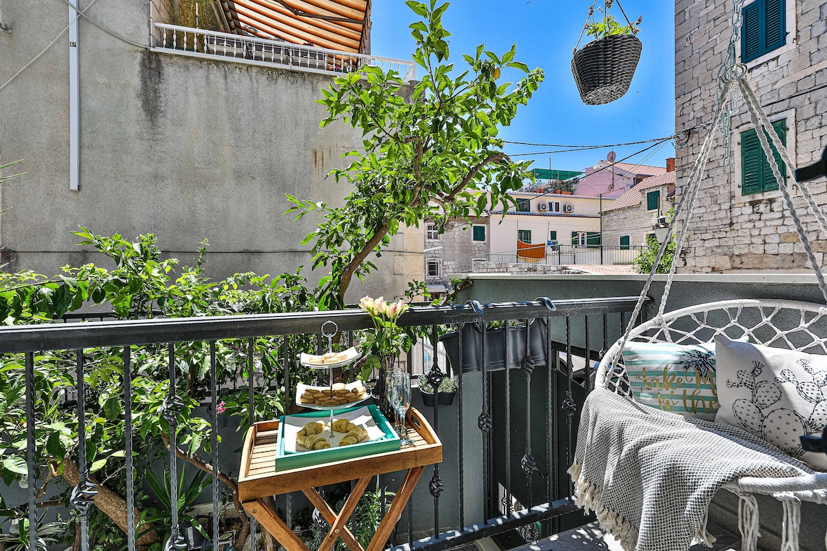 A balcony area is visible, featuring greenery including a tree and hanging plants. A cozy swing chair is arranged beside a small wooden table, which holds treats on a tray. Sunlight brightens the space, creating an inviting outdoor atmosphere.