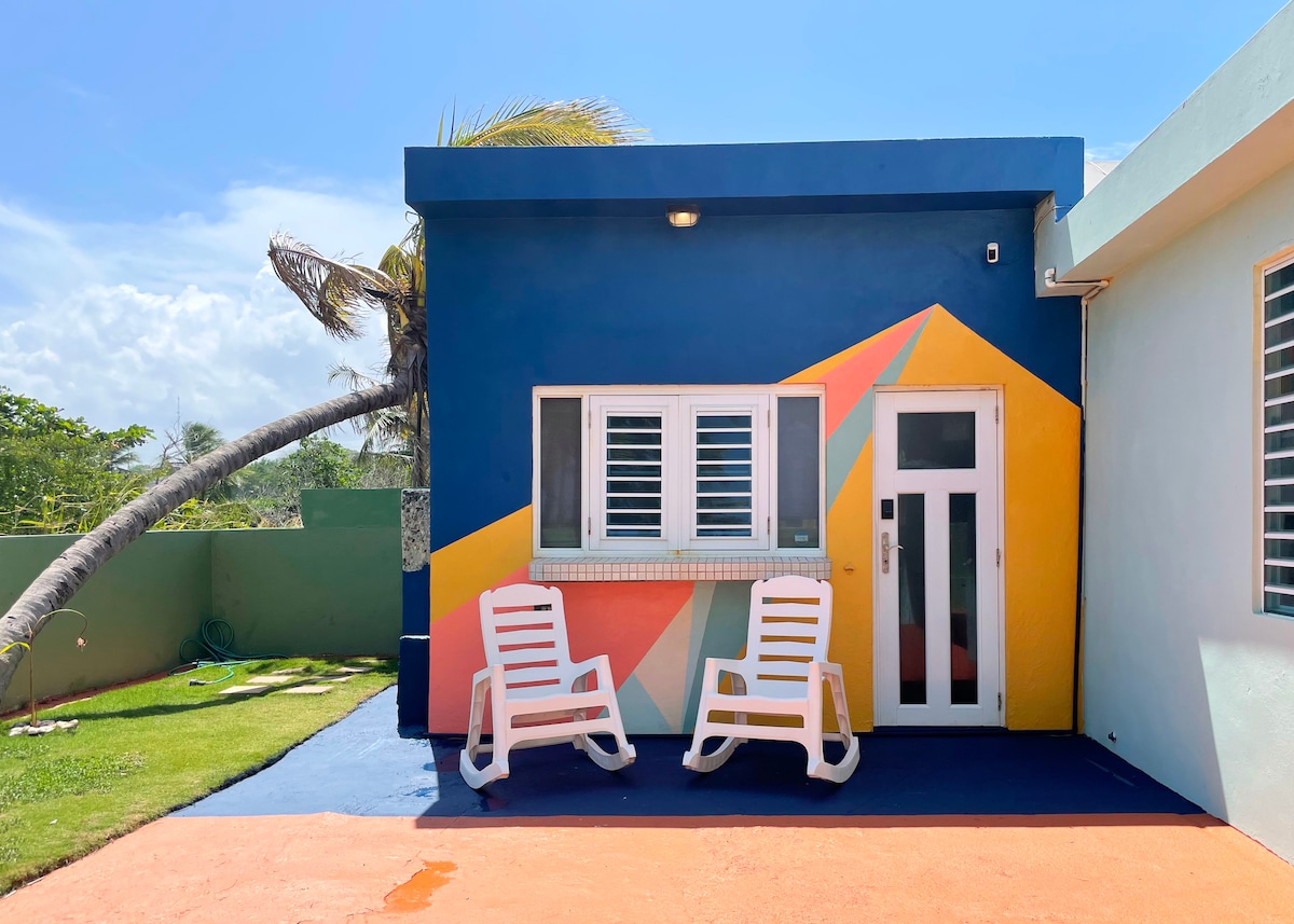 The exterior of the bungalow features a vibrant mural with geometric shapes in colorful hues. Two white lounge chairs are positioned on a painted patio surface, inviting relaxation. Lush green grass is present, complemented by tropical palm trees in the background, all under a clear blue sky.