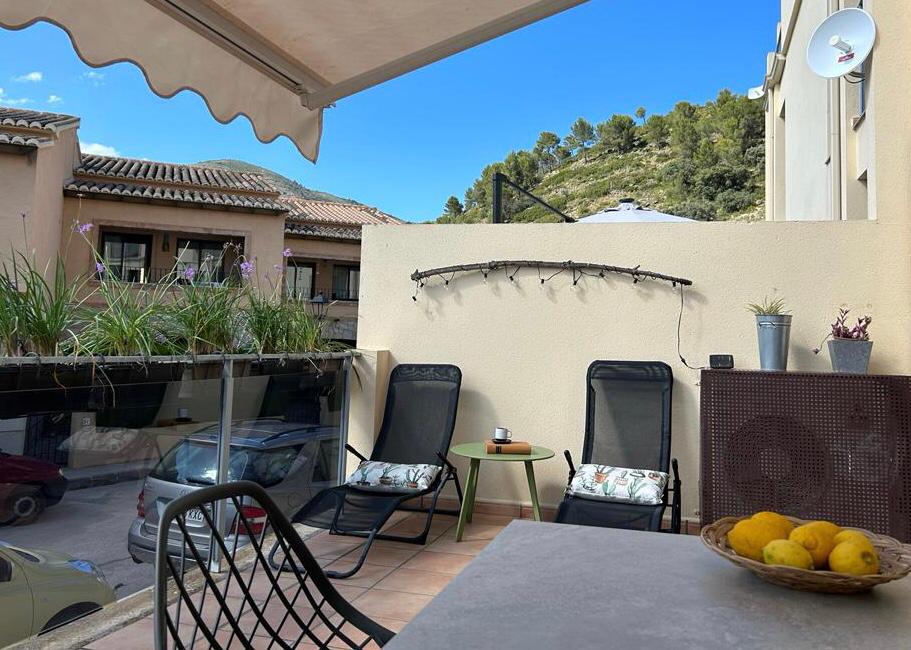 A balcony space is presented with two lounge chairs positioned for relaxation. A small table sits nearby, and a bowl of lemons adds a pop of color. Green plants line the railing, complementing the serene view of the surrounding hills under a clear blue sky.