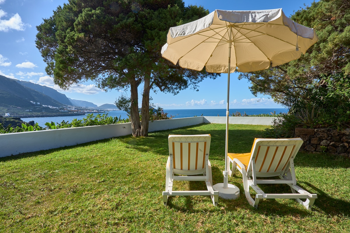 Two lounge chairs with cushions are positioned under a large umbrella on a grassy area, offering views of the sea and surrounding mountains. The landscape features trees and a clear sky with fluffy clouds, providing a tranquil outdoor space for relaxation.