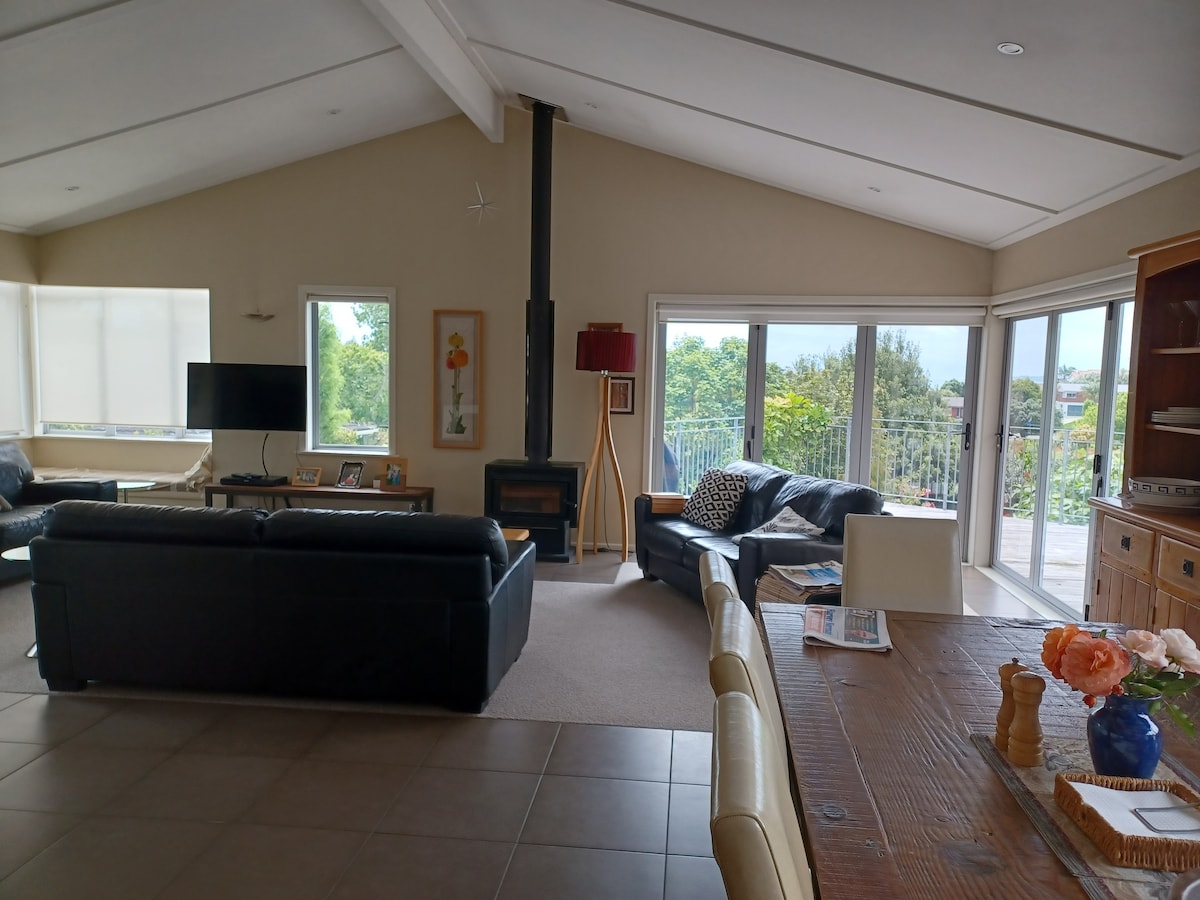 A spacious living area is presented with large glass doors offering distant views of greenery and the sea. Comfortable sofas face a modern woodburning stove, and a dining area is adjacent, featuring a wooden table adorned with fresh flowers. Natural light fills the room.