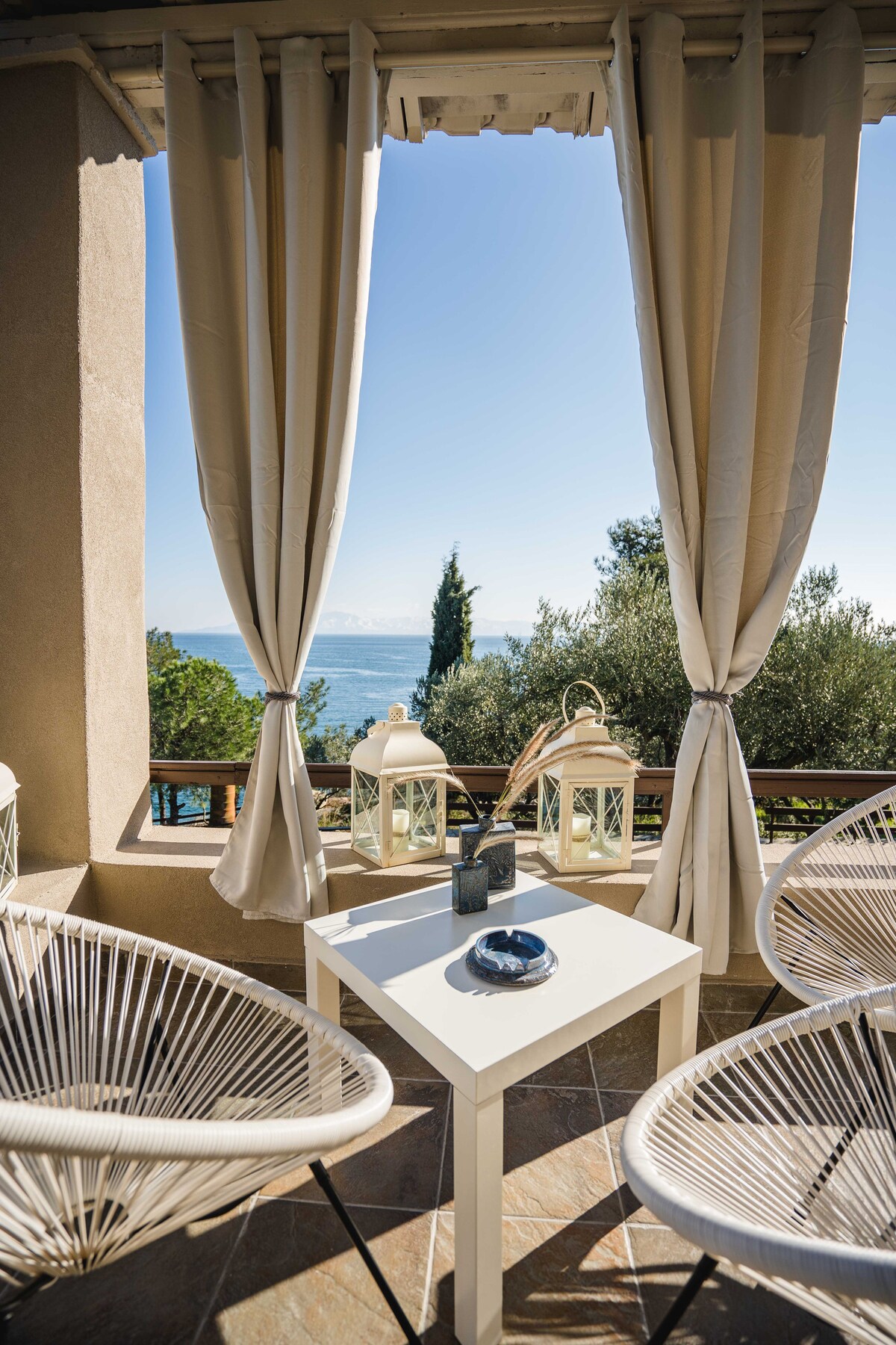 An inviting outdoor seating area features two stylish chairs and a small white table. Soft curtains frame the space, providing a sense of privacy while revealing views of the Aegean Sea and surrounding greenery. Natural light enhances the serene atmosphere.