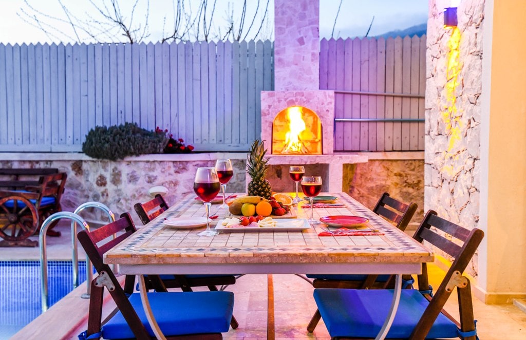 A dining area is presented with a large table set for a meal, featuring vibrant glasses of red wine and an assortment of colorful fruits. A stone barbecue is visible in the background, alongside a pool, and warm lighting enhances the inviting ambiance.
