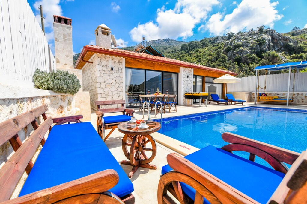 A private swimming pool is surrounded by wooden loungers with blue cushions. A stone-built barbeque and dining area are visible under a shaded pergola. Lush greenery and mountains provide a scenic backdrop, while large glass doors connect the outdoor space to the interior.