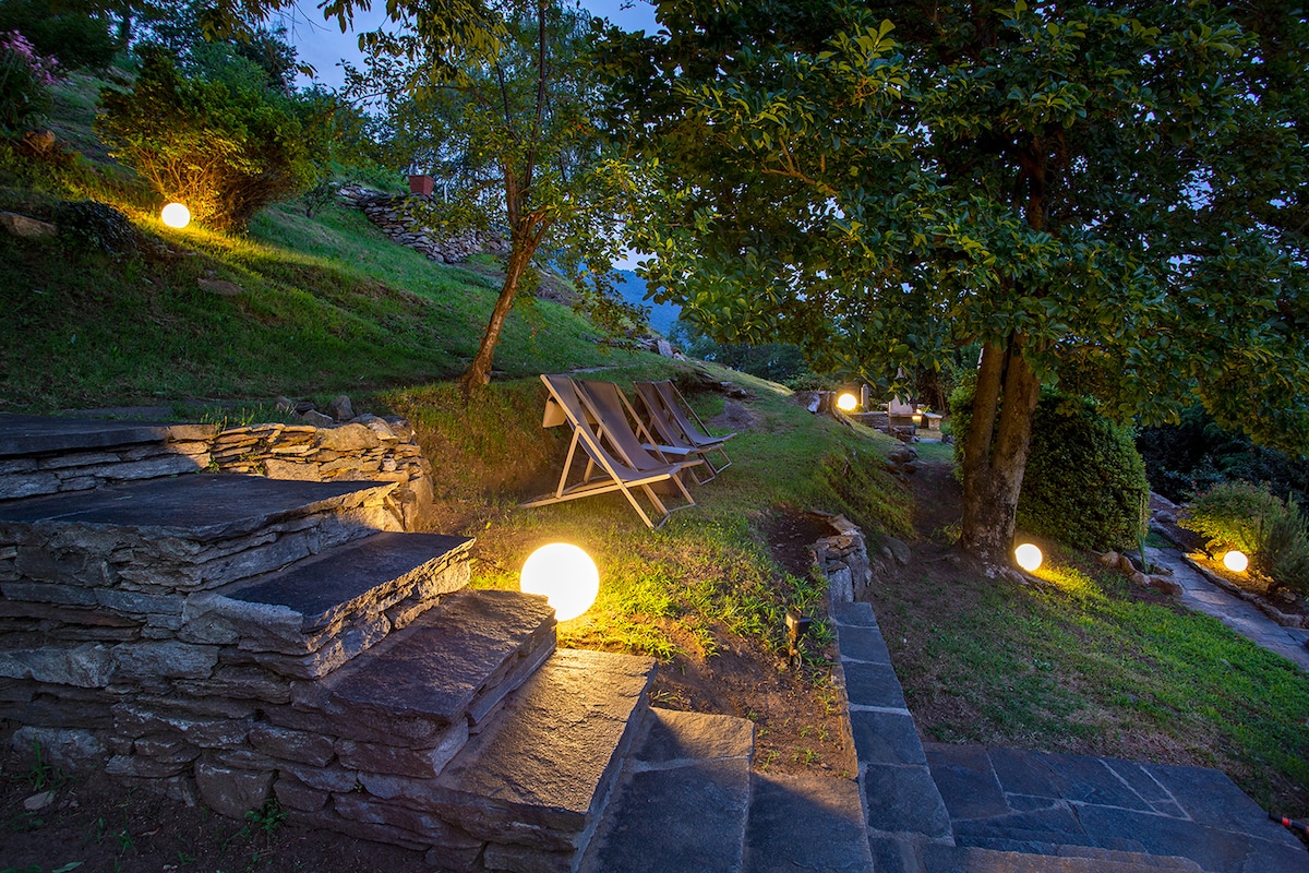 The garden features gently sloping paths lined with smooth stone steps and illuminated by soft light globes. Comfortable lounge chairs are positioned on the grassy area, surrounded by lush greenery and trees, creating a serene outdoor retreat.