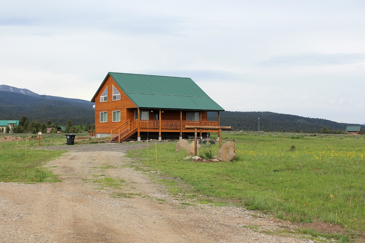 A newly constructed cabin is shown surrounded by an open grassy area, featuring a green metal roof and wooden exterior. A wide front porch with railing adds to its inviting design. Scenic mountains are visible in the distance under a cloudy sky.