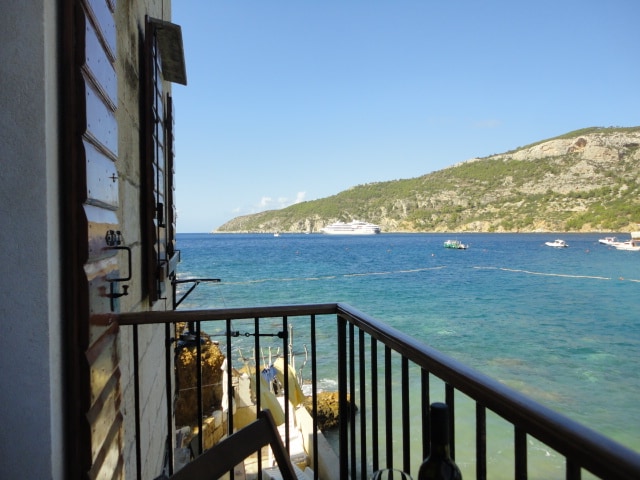 A scenic view from a balcony overlooking a calm sea, with small boats visible in the water. The lush green hillside can be seen in the distance, complementing the clear blue sky. The balcony features a railing with a rustic design.