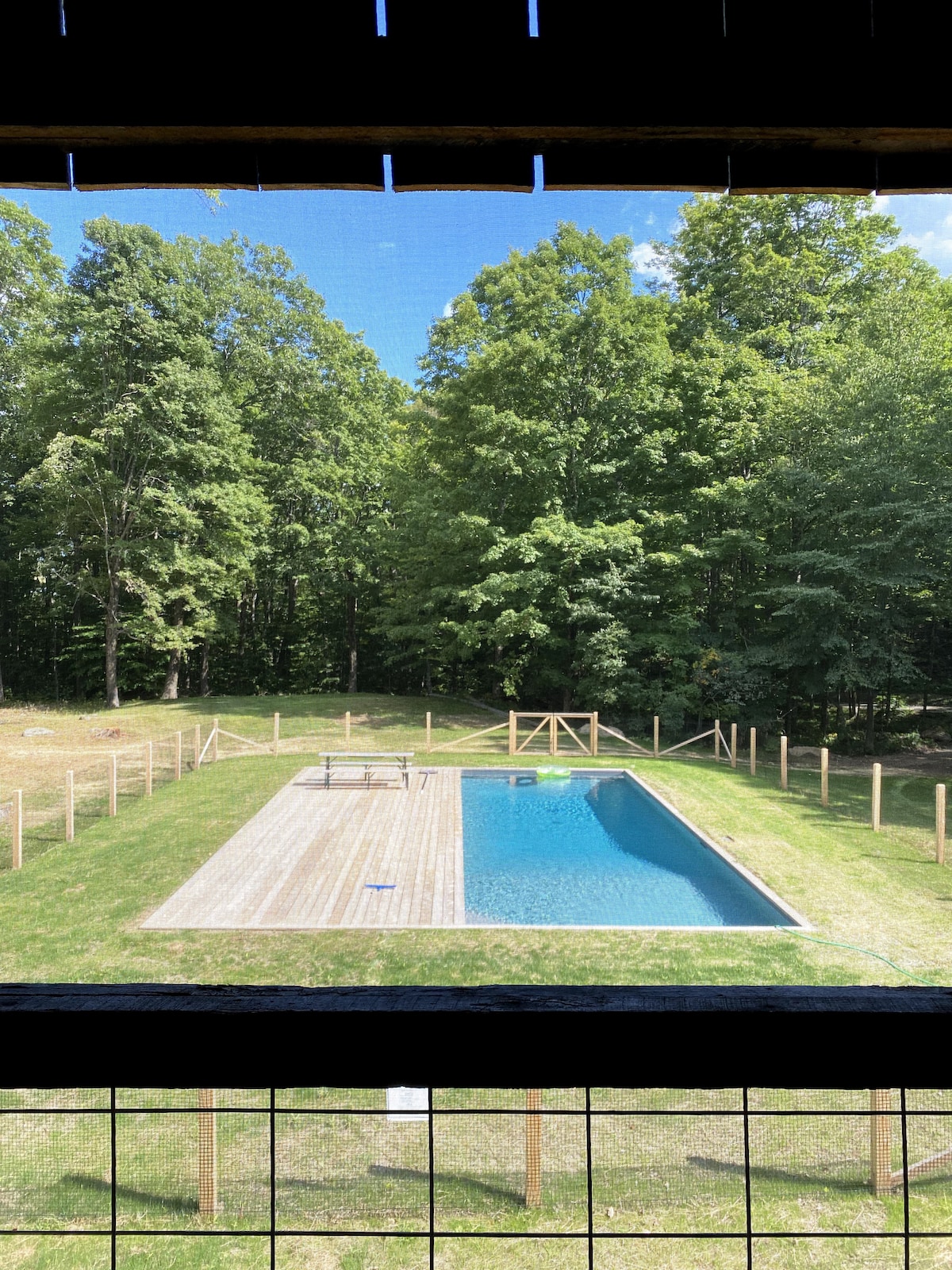 A serene outdoor space is depicted, featuring a rectangular swimming pool surrounded by a deck. Lush green trees create a natural backdrop, enhancing the tranquil ambiance. A fenced area offers privacy and ample lawn space for relaxation and leisure activities.