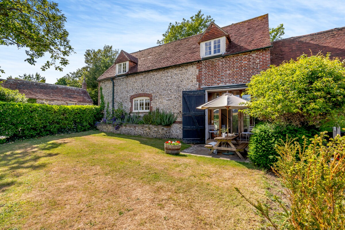 The cottage's exterior features a charming combination of flint and brick. A large garden area surrounds the property, with neatly trimmed hedges and vibrant greenery. A wooden picnic table is covered by an umbrella, providing shaded seating for outdoor dining.