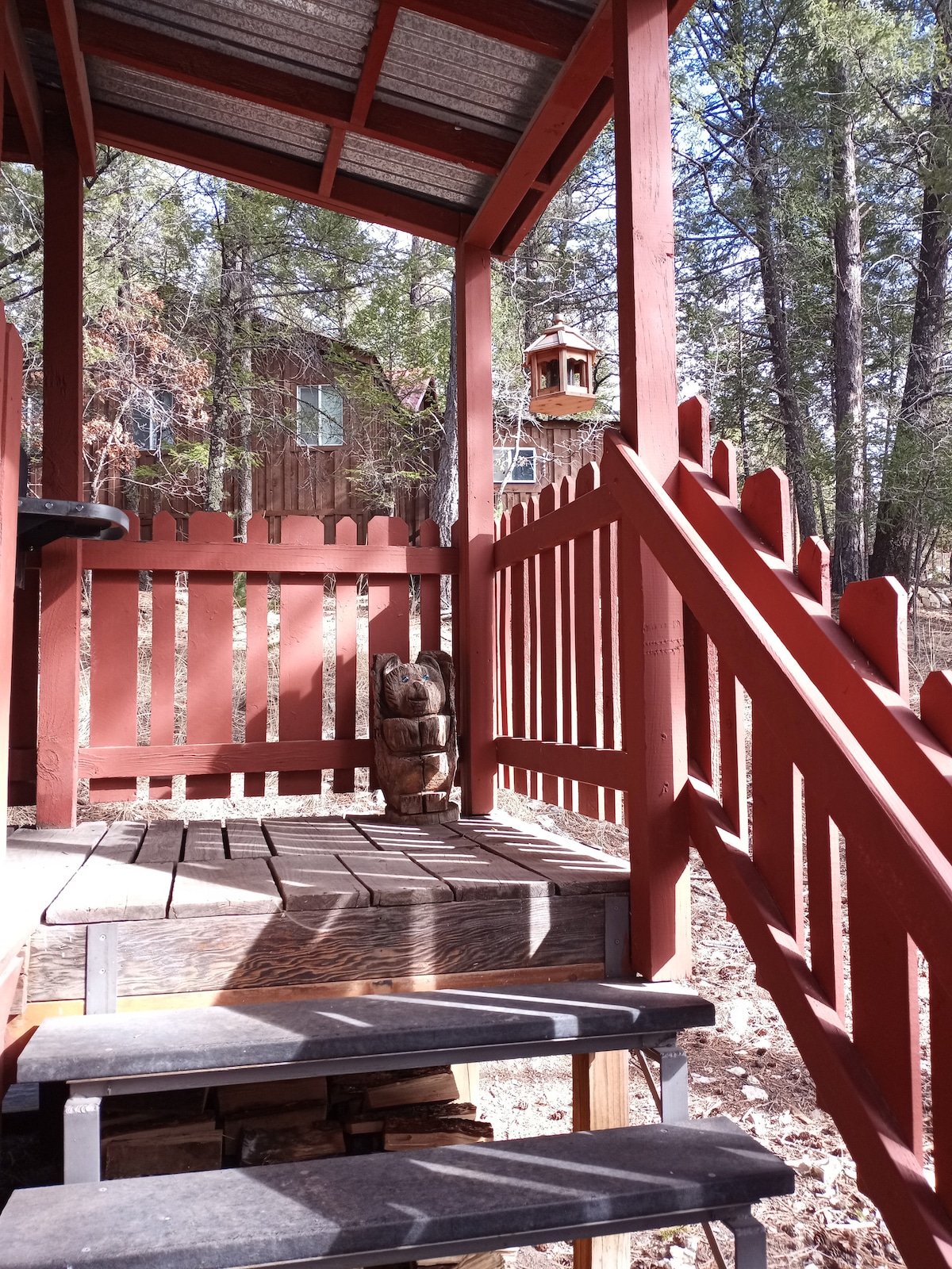 Wooden steps lead to an outdoor porch area, enclosed by a red picket fence. A carved wooden bear adds charm to the setting, surrounded by tall trees that create a natural backdrop.