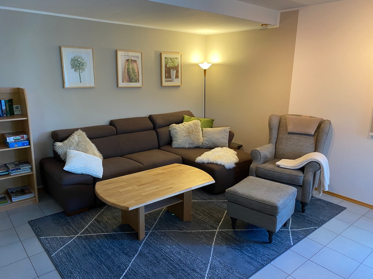A cozy living area features a sectional sofa adorned with soft throw pillows and blankets. A wooden coffee table is positioned on a patterned rug, while framed botanical prints hang on the wall. A standing lamp provides warm lighting, enhancing the room's comfort.