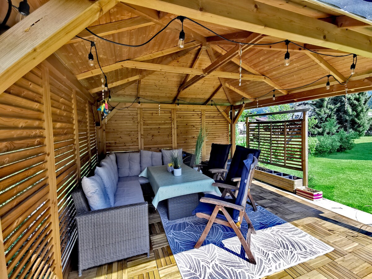 A shaded outdoor pavilion features a comfortable seating arrangement with plush cushions and a central table. Soft lighting hangs overhead, illuminating the space. An area rug adds texture underfoot, while greenery can be seen in the background, creating a serene outdoor environment.