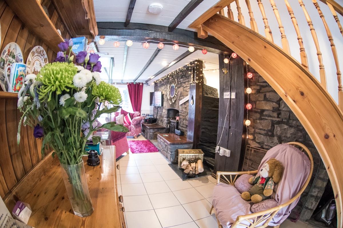 A cozy living area features a stone wall with a log fire, accented by colourful fairy lights. A bouquet of fresh flowers is displayed on a wooden console, while a plush chair adds comfort. Soft red curtains frame windows, inviting light into the space.